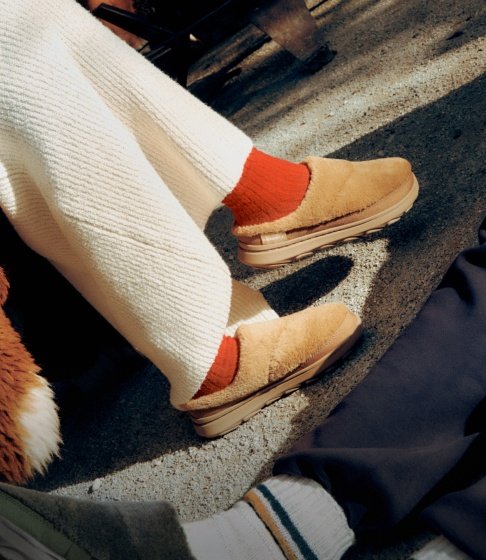 Person in tan Merrell slip-on shoes, orange socks and cream knit trousers relaxing outdoors.