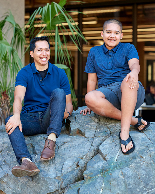 A big brother sitting on a boulder with a little brother smiling.