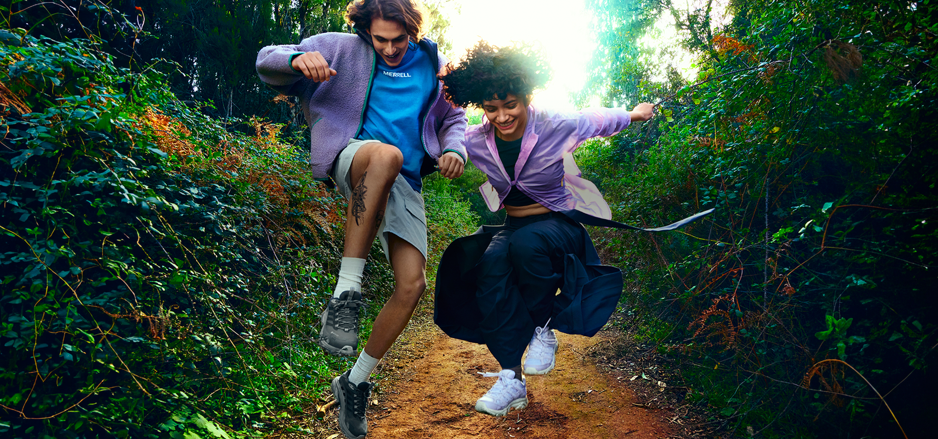 Two people jumping on a dirt path wearing Merrell hiking shoes.