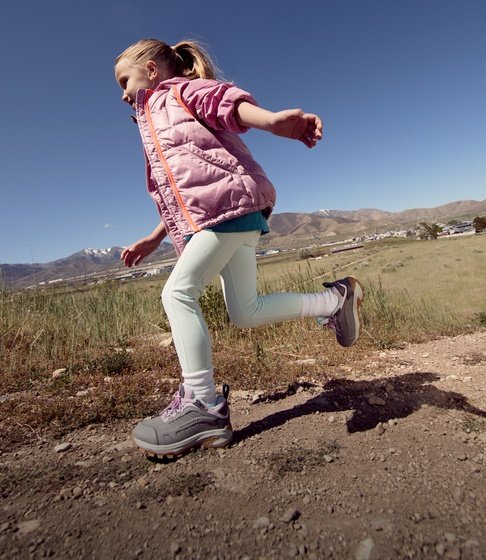 Girl in a pink vest running along a dirt trail with mountains in the background.