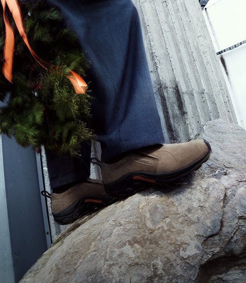 Brown Merrell slip-on shoes beside an evergreen wreath tied with an orange ribbon.