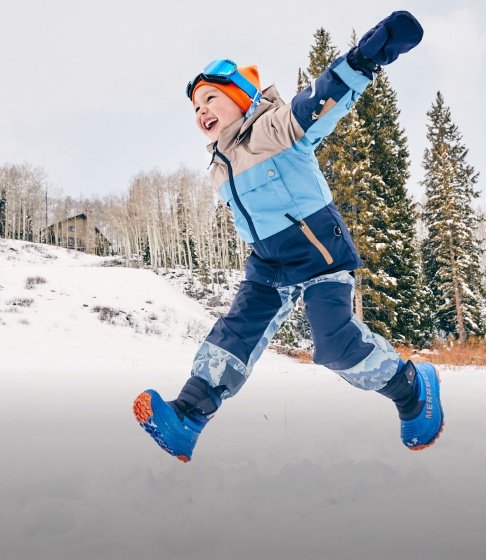 Garçon riant en habit de neige bleu, lunettes de ski et bottes d’hiver Merrell bleues sautant dans la neige, avec des sapins derrière lui.