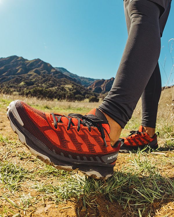 Les pieds d'une personne portant des chaussures Merrell dans un champ avec des montagnes en arrière-plan.