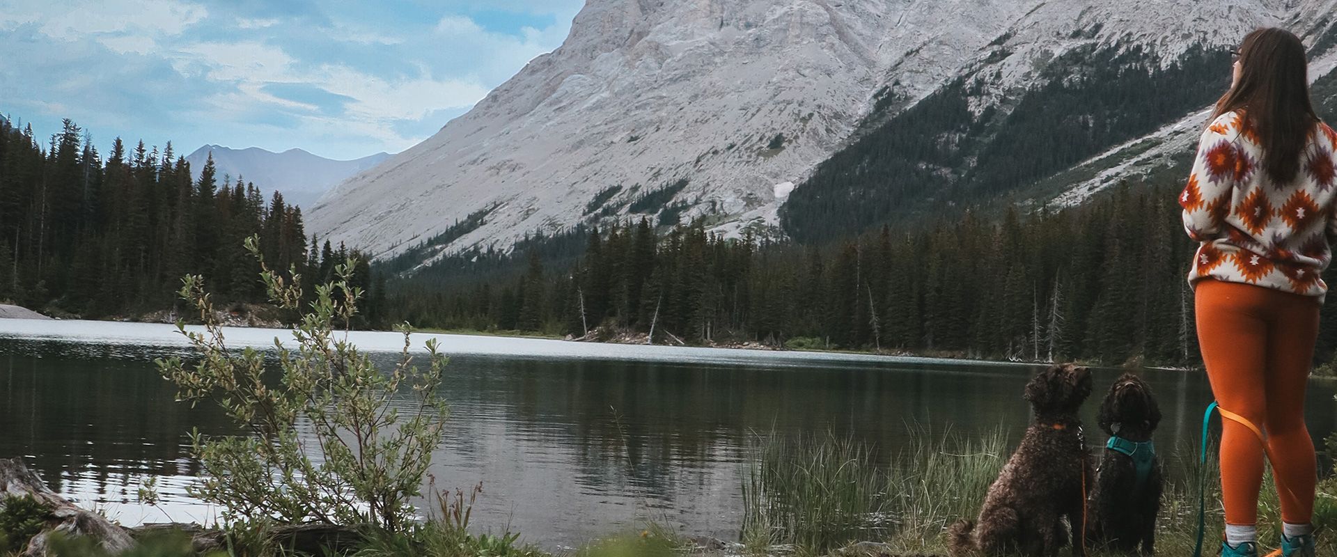 Janine Dersch avec deux chiens près d'un lac avec des arbres et des montagnes en arrière-plan.