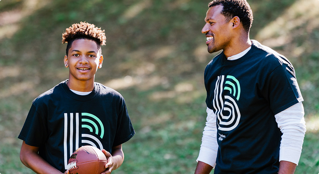 A little brother smiling, holding a football, with a big brother smiling.