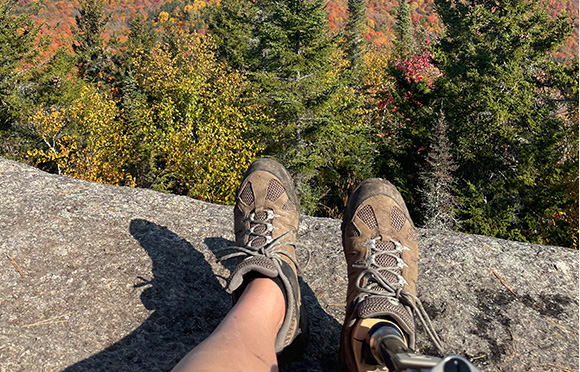 A person's legs and Merrell hiking shoes on a rock with trees in the background.