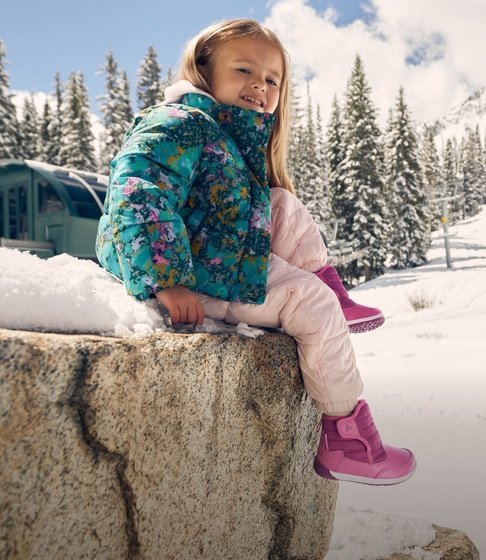 Girl in a floral puffer jacket and pink winter boots sitting on a snowy boulder with snow-covered pine trees behind her.