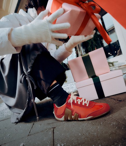 Person in red Merrell shoes holding a wrapped gift beside a stack of presents.