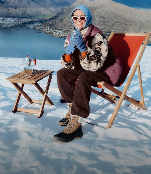 Person in tan Merrell winter boots sitting in a deck chair in the snow above a mountain lake.