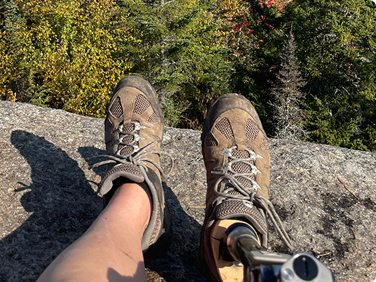 A person's legs and Merrell hiking shoes on a rock with trees in the background.