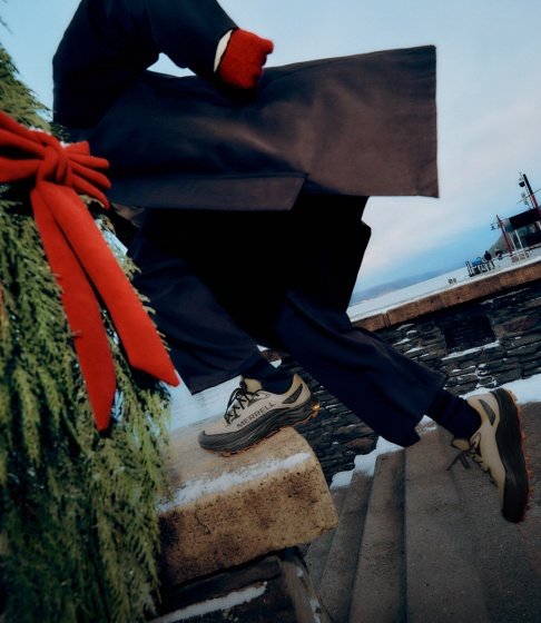 Person in a dark coat and tan Merrell shoes walking up snowy stone steps beside holiday greenery tied with a red bow.