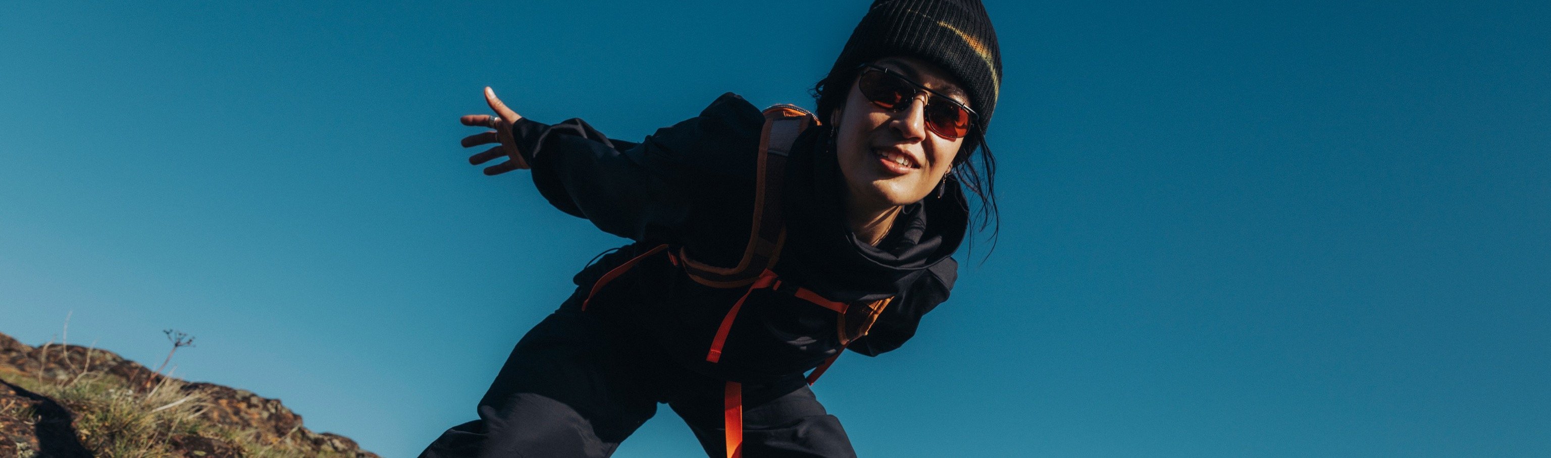 Hiker in a black beanie and jacket leaning over rocky terrain under a blue sky