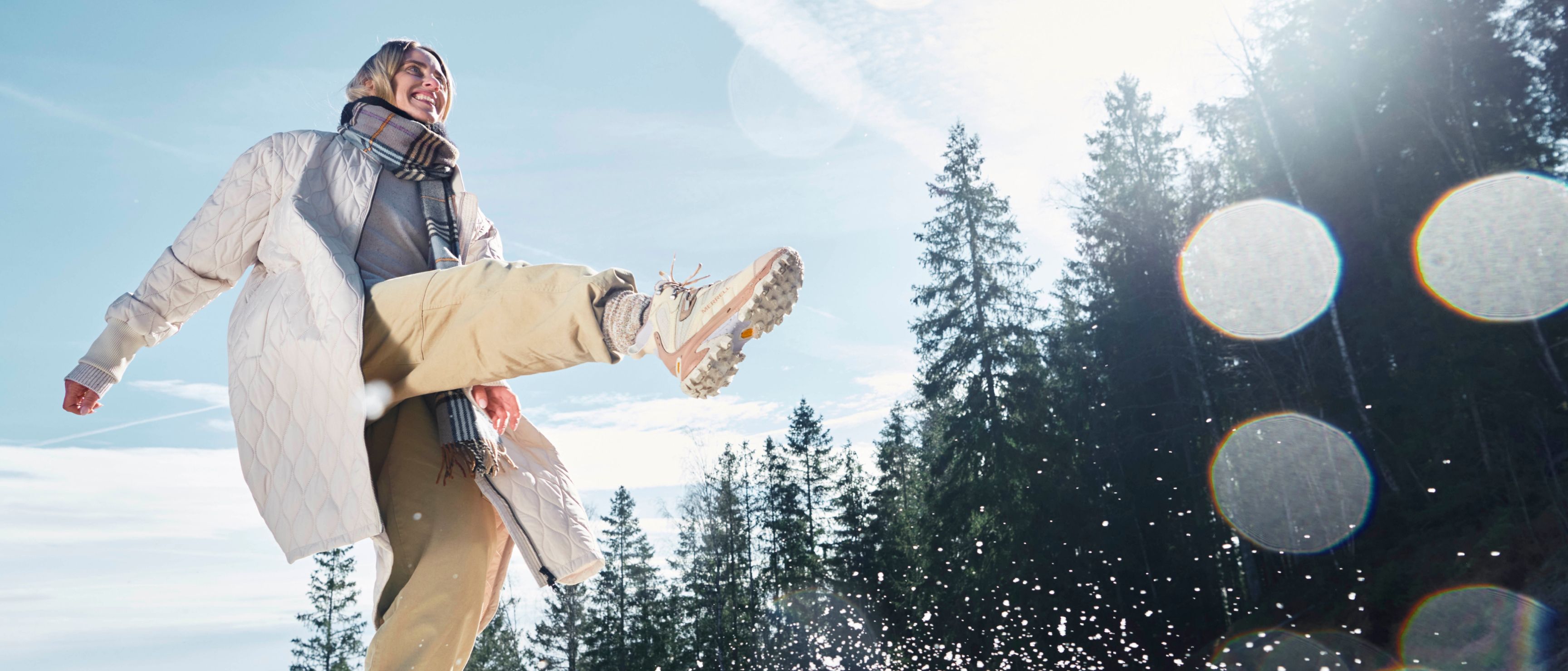 Person wearing Merrell winter gear jumping in the snow with pine trees in the background.