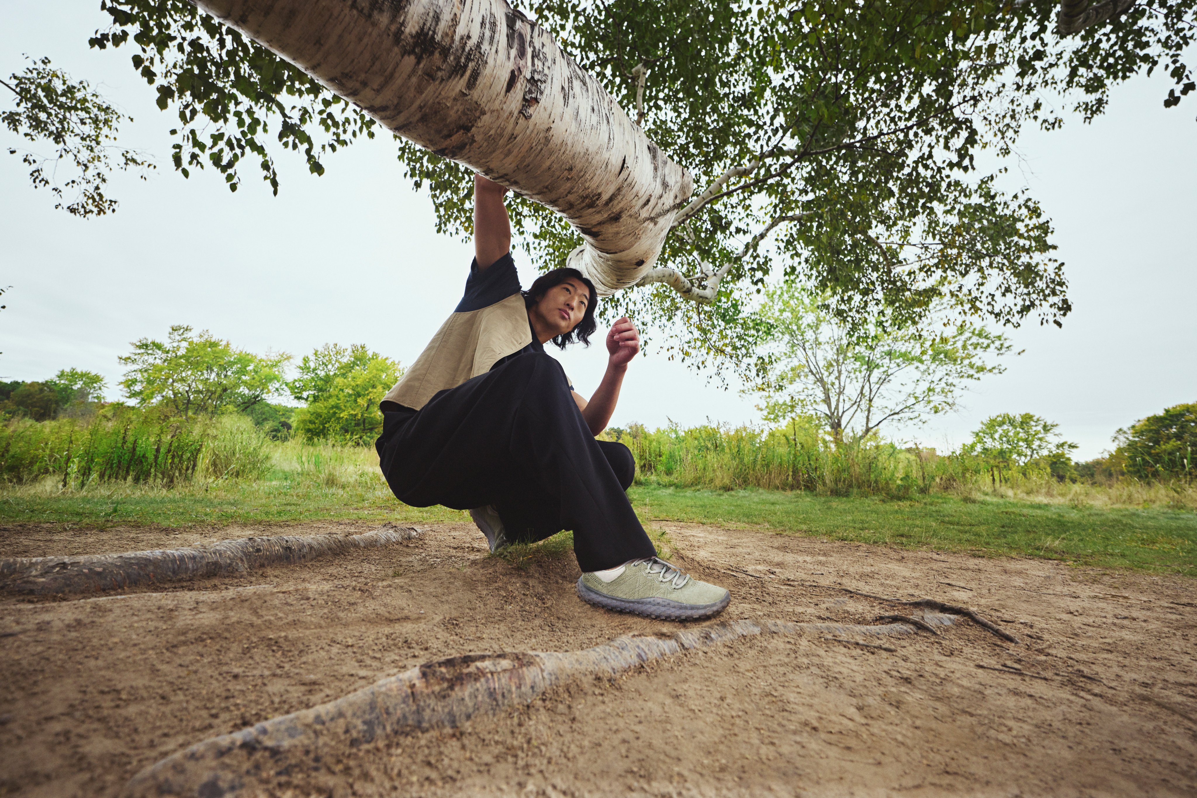 A man wearing Merrell shoes crouching beneath a birch tree branch.