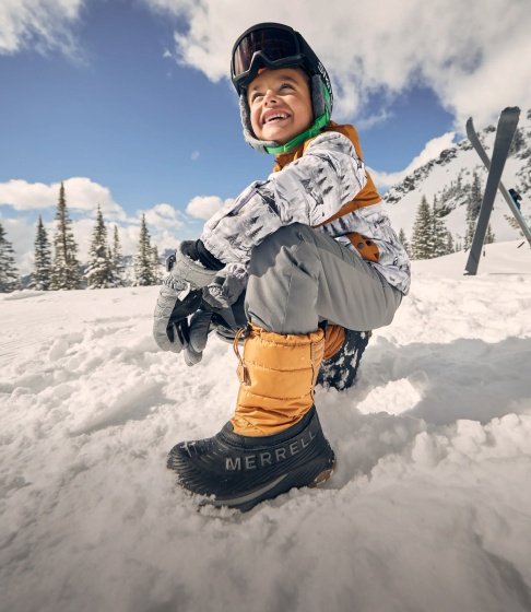 Smiling boy in a green ski helmet and goggles crouching in the snow in orange Merrell winter boots, with skis and snowy pine trees behind him.