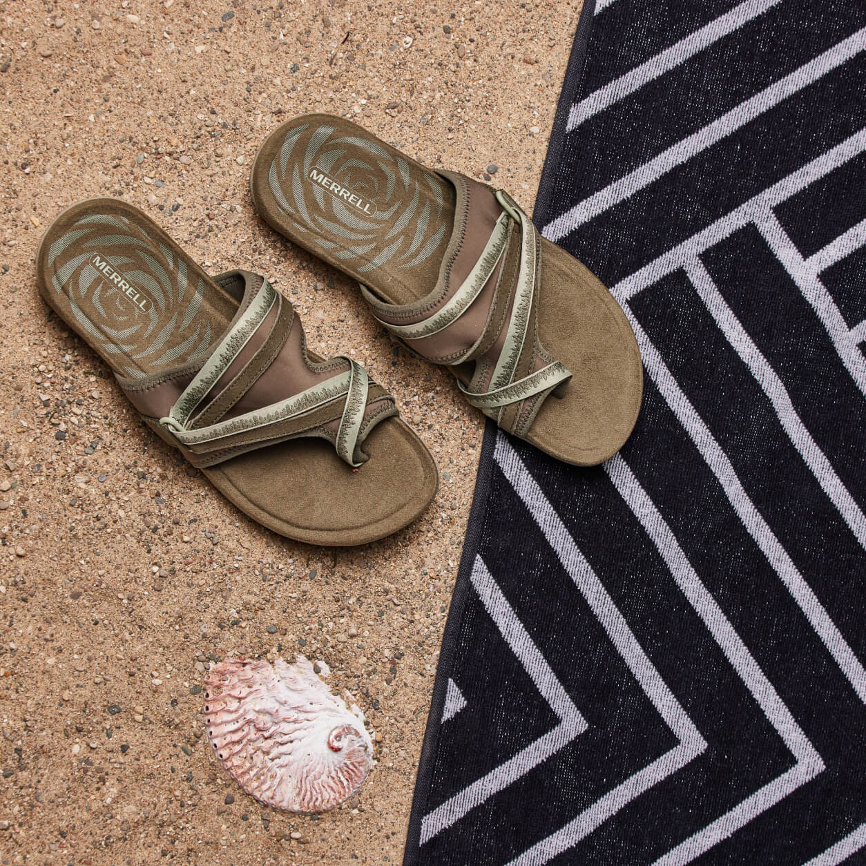 Sandals at the beach, next to a sea shell and a beach towel.