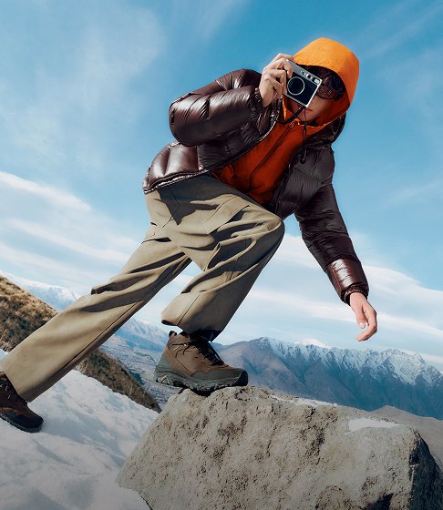 Person in an orange hood and Merrell hiking boots taking a photograph on a snowy mountain.