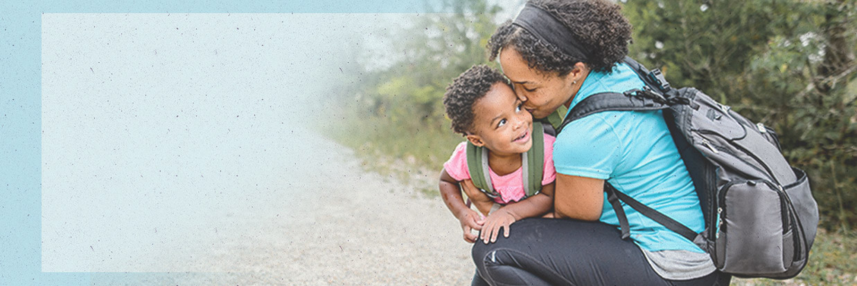 Mother wearing backpack is squatting down to hug her child. Both are smiling and wearing Merrell hiking shoes.