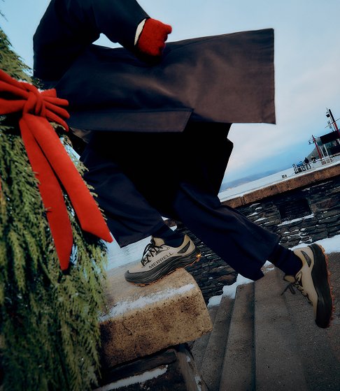 Person carrying a wrapped gift up snowy stone steps in Merrell hiking shoes beside holiday greenery.