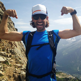 A dark-haired man in a baseball cap and a bright blue t-shirt grins and flexes both biceps in front of a mountain vista.