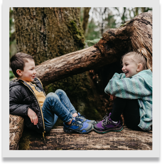 Two kids talking and laughing, sitting at the base of a tree in the woods.