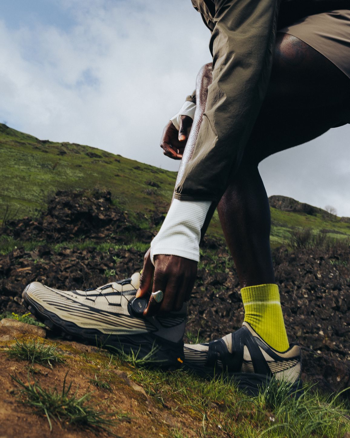 A trail runner adjusting a Merrell shoe on a rocky hillside.