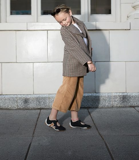 A blonde-haired girl with sunglasses who's about 10 years old looks down at her shoes while standing on the sidewalk next to a white tiled building with pillars