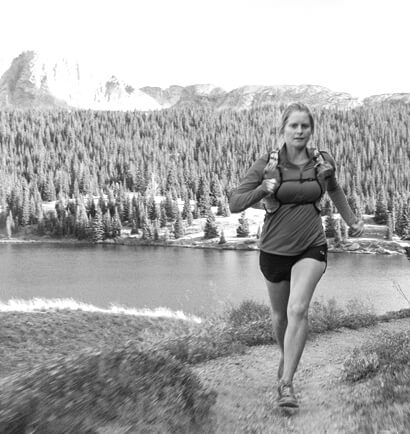A runner rounds a corner on a trail. Behind and below her is a lake, and behind that a thick forest and a mountain.