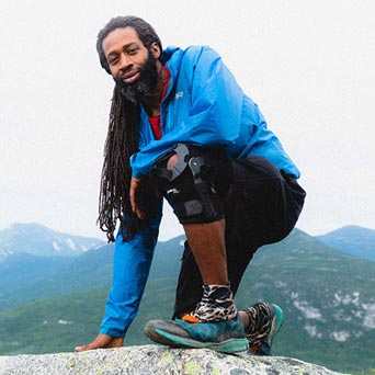 A long-haired man in a bright blue shirt grins in front of a mountain vista.