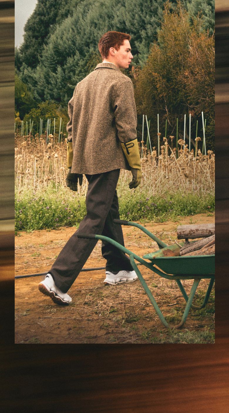 Person in a tweed jacket pulling a wheelbarrow of firewood across a field.
