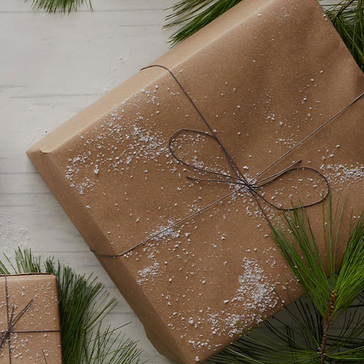A brown paper-wrapped present with snow sprinkled on it, and green pine bows in the background.
