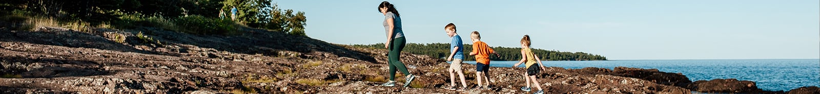 An adult hiking with 3 kids by a lake.