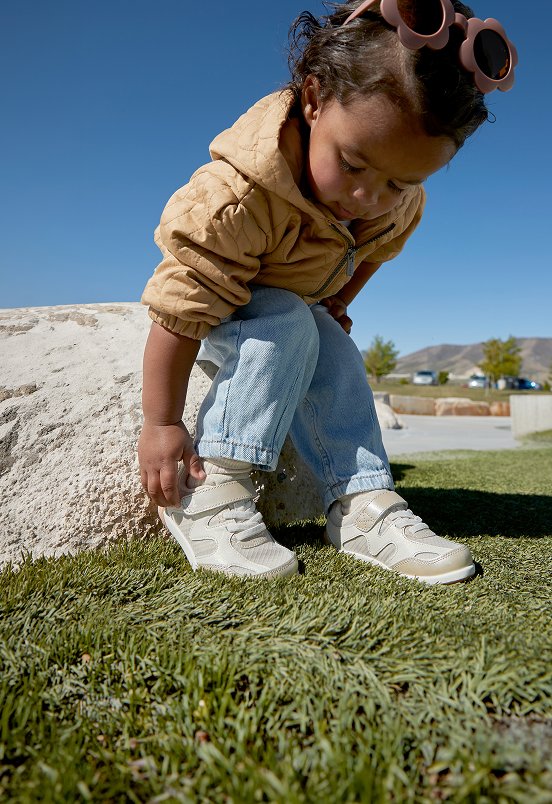 Child sitting on a rock in the grass, adjusting the strap on their Merrell sneakers.