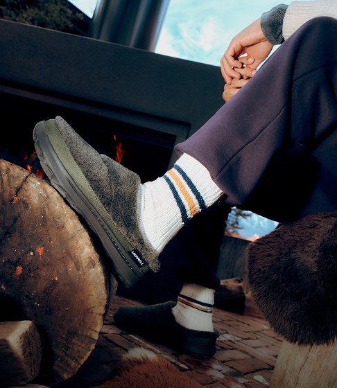 Person in grey Merrell slip-on shoes and striped socks resting their feet beside a wood-burning stove.
