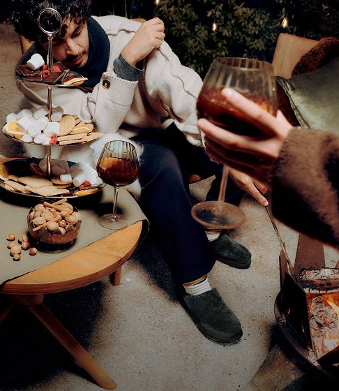 Friends in cosy Merrell slip-on shoes sharing drinks and snacks beside a wood-burning stove.