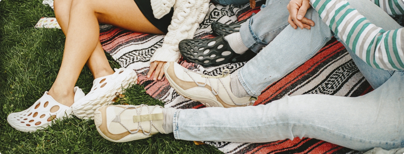 Legs of a three people sitting on a blanket in the grass wearing Merrell shoes.