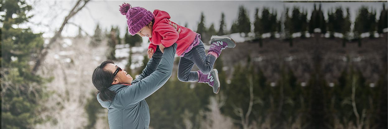 Mother lifting up her child outside in the snow. Both are smiling and wearing Merrell hiking shoes.