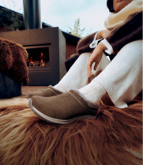 Person in brown Merrell slip-on shoes and white socks relaxing beside a wood-burning stove.