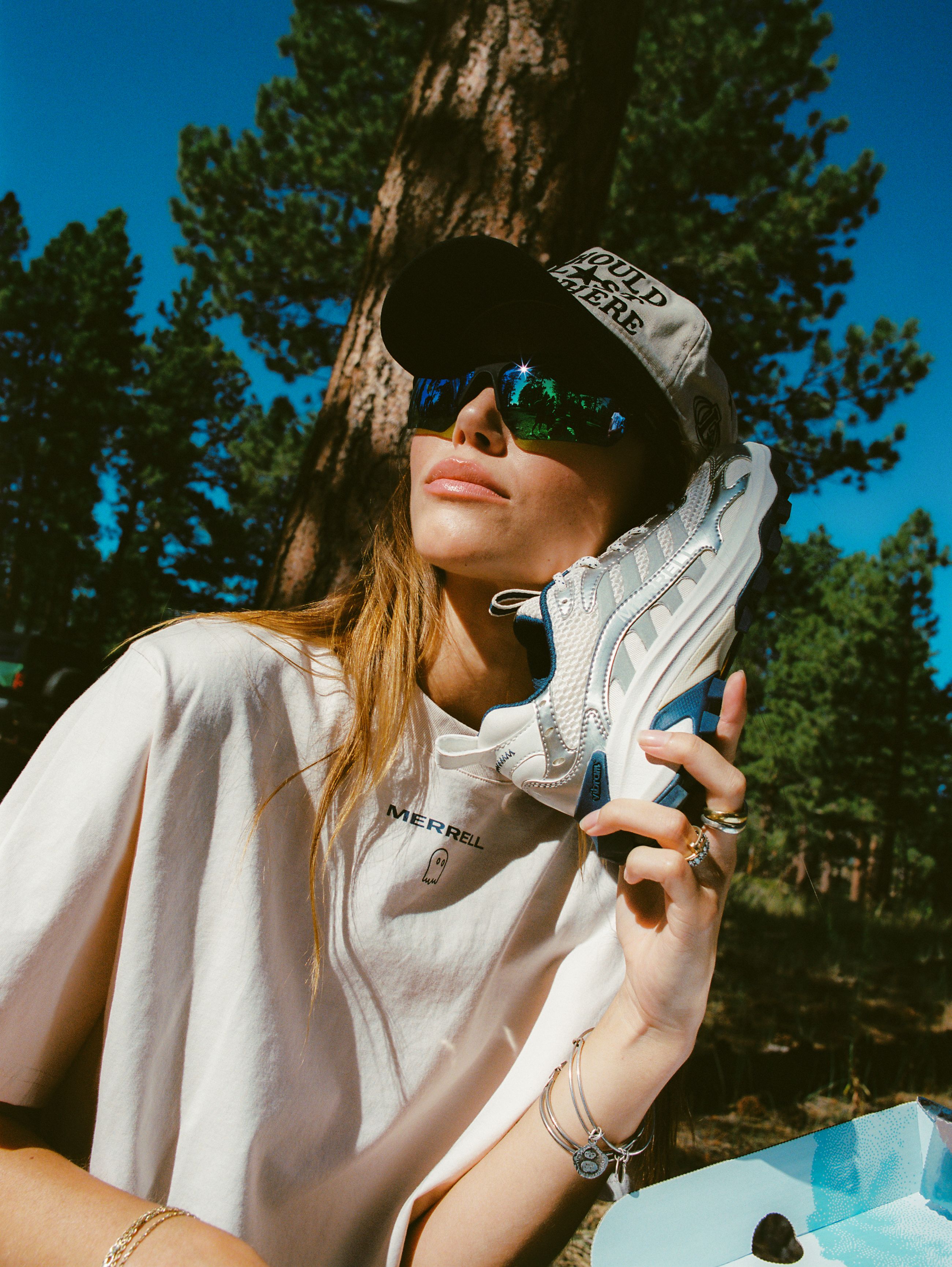 Person looking cool wearing a Lonely Ghost hat, sunglasses, and a Merrell shirt holding a Merrell shoe next to their face.