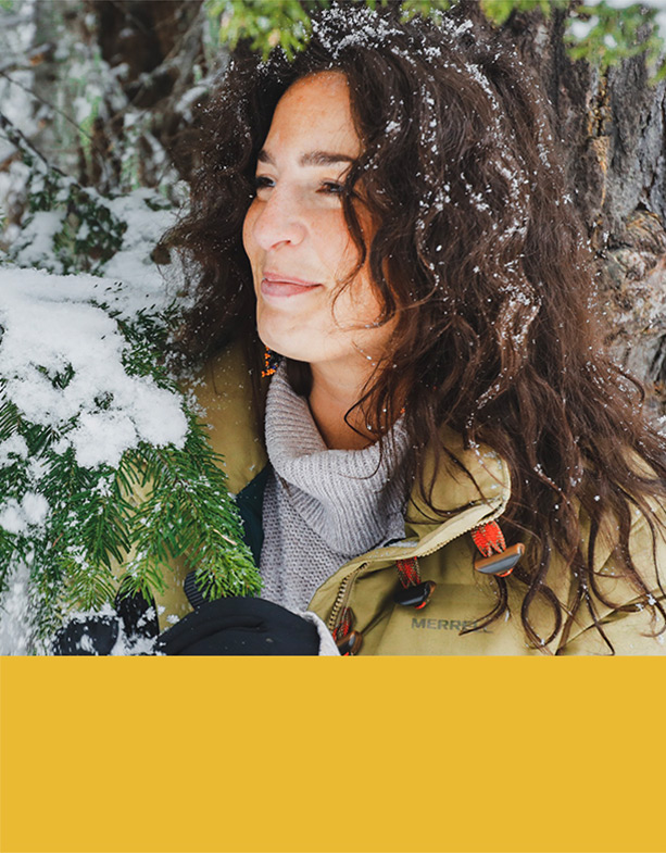 Woman standing outside in the snow by a pine tree.